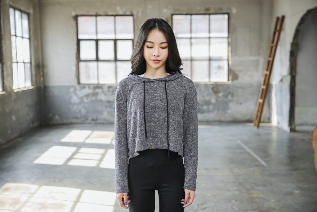 A woman in trendy athleisure attire, donning a gray sweater and black pants, stands confidently in an empty room.