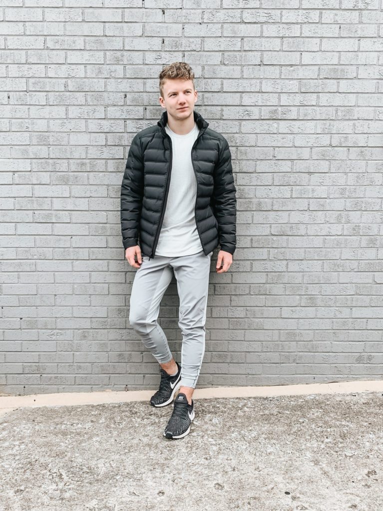 A man, embracing the athleisure trend, leans against a brick wall donning a black puffer jacket.