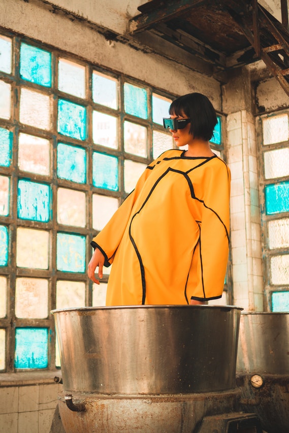 A woman in a yellow coat showcasing her clothing collection in front of a large pot.