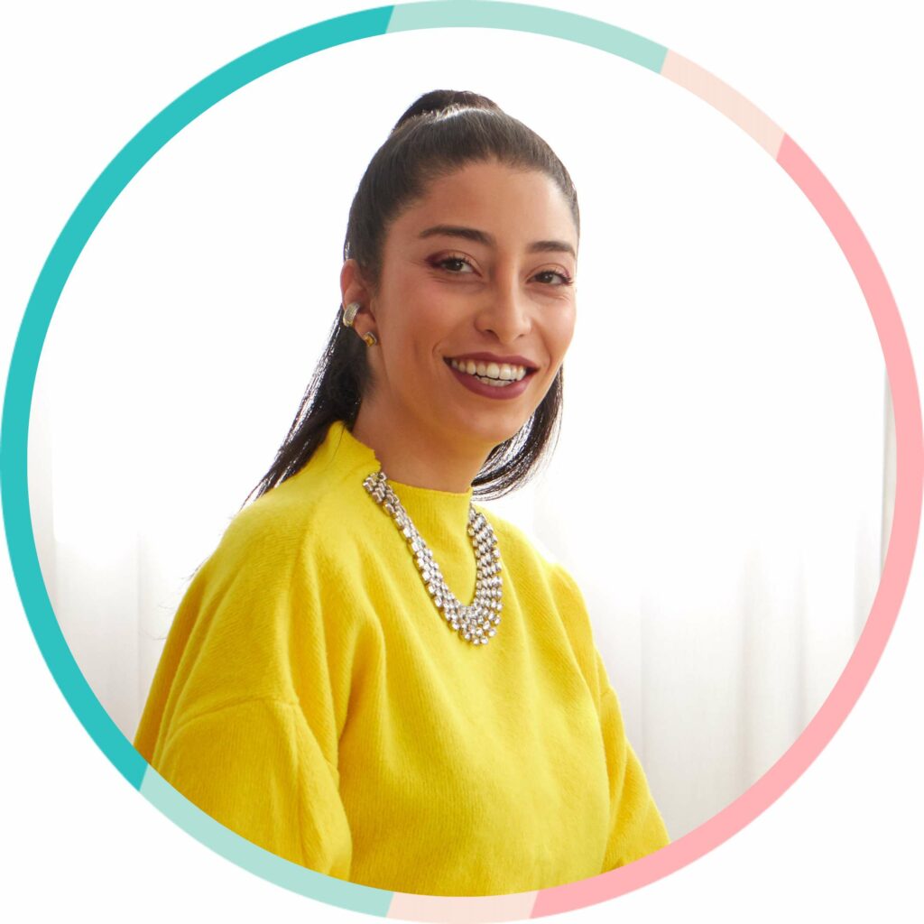 A woman in a yellow sweater smiling in front of a circle, showcasing her fashionable style and confidence.