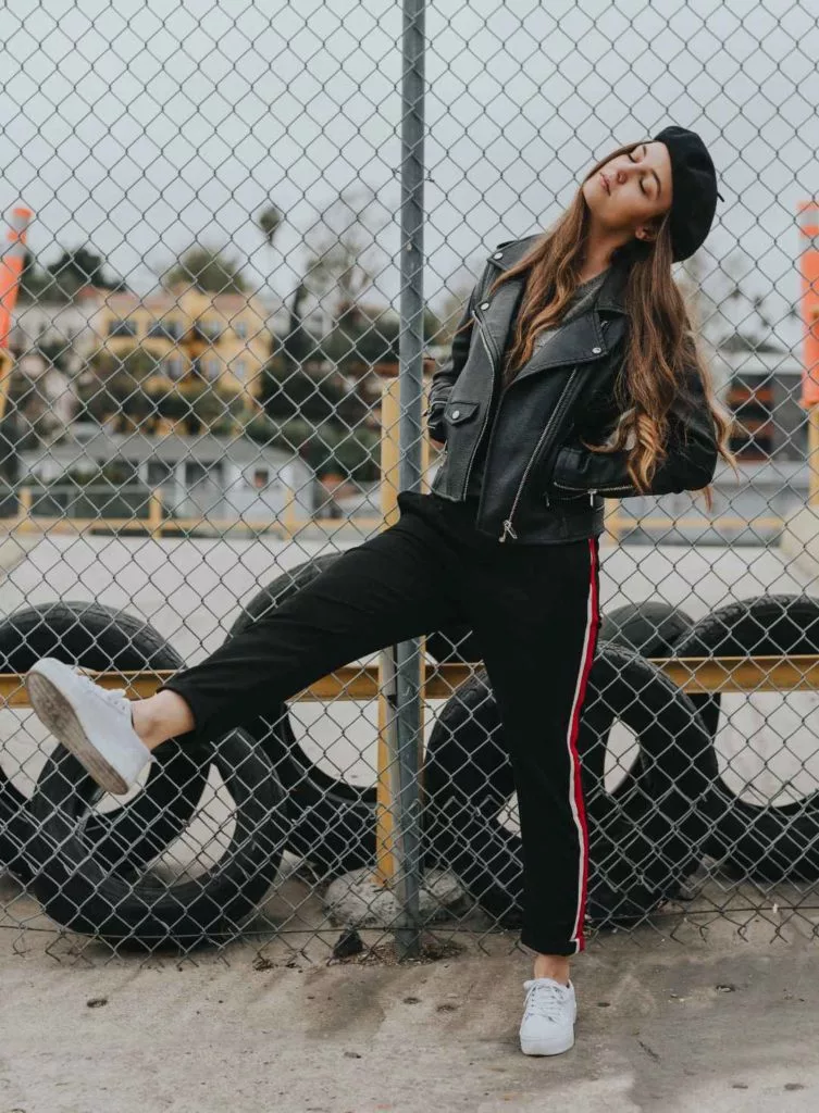 A woman leaning against a chain link fence in casual wear - black track pants and a black leather jacket.
