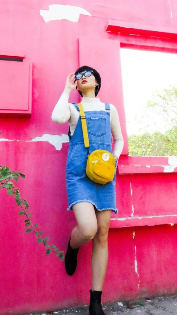 A casual woman in overalls leaning against a pink wall.