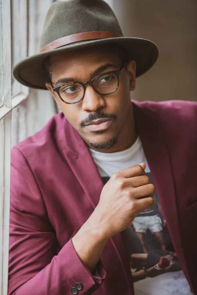 A black man in casual wear, wearing a hat and glasses, leaning against a window.